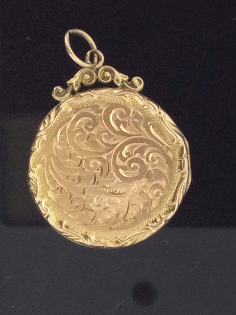 May include: A gold locket with an ornate, engraved design. The locket has a loop at the top for attaching a chain.