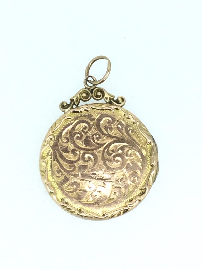May include: A gold locket with an ornate, engraved design. The locket has a round shape and a bail with a decorative loop.