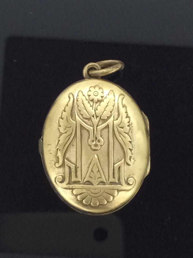 K&ouml;nnte beinhalten: Ein ovales goldenes Medaillon mit einem gravierten floralen Design und dem Buchstaben "M".