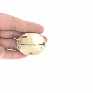 May include: A gold-toned oval brooch with a textured surface and a pin on the back.