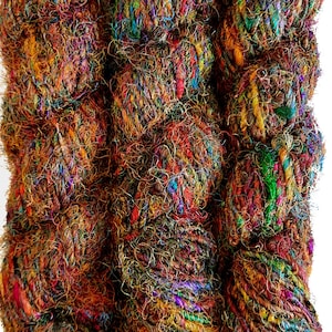 以下が含まれることがあります： カラフルで多様な質感の糸が3玉。糸は茶色、オレンジ、赤、緑、青、紫のミックスです。糸はリサイクルされたサリーシルクで作られています。