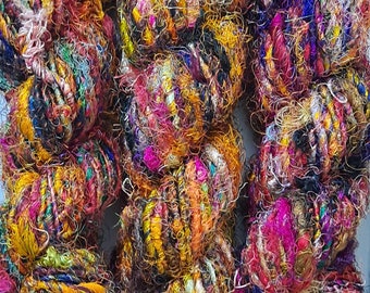 May include: Three skeins of colorful yarn, each a mix of textures and colors, including gold, pink, purple, blue, and green. The yarn is made of silk and other fibers.