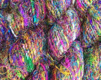 Könnte beinhalten: Nahaufnahme eines Stapels bunter Wolle, mit Farbtönen von Lila, Blau, Grün, Gelb und Orange. Die Wolle besteht aus recycelten Materialien und hat eine einzigartige, strukturierte Optik.