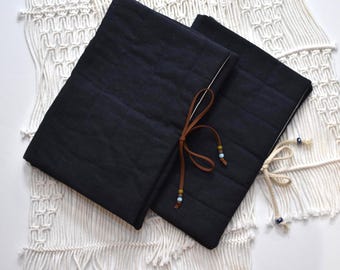 May include: Two dark blue quilted fabric journal covers with a brown leather tie and decorative beads. The covers are rectangular and have a stitched grid pattern. The background is a white macrame textile.