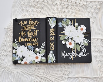 May include: A black journal with a gold inscription that reads "We love because He first loved us. 1 John 4:19". The journal is decorated with white flowers and gold accents. The date "Oct. 21, 2023" and the name "Kuczkowski" are also written in gold on the cover.