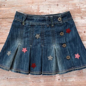 May include: A denim skirt with a pleated design and button closure. The skirt is adorned with red and black checkered star-shaped patches. The skirt has a classic, casual style.