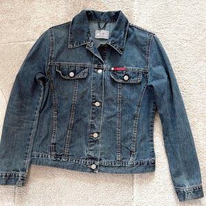 Puede incluir: Chaqueta vaquera azul oscuro con cierre de botones, dos bolsillos en el pecho y cuello. La chaqueta tiene un diseño clásico con botones plateados y un pequeño logotipo rojo y blanco en el bolsillo del pecho.