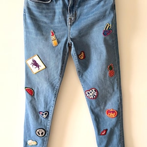 Peut inclure: Jean bleu clair orné de divers écussons colorés. Les écussons comprennent un rouge à lèvres, un taureau, une pastèque, un ananas et des motifs en forme de cœur. Le jean est présenté sur un cintre.