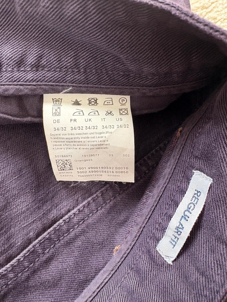 Puede incluir: Primer plano de unos vaqueros morados con una etiqueta que muestra las instrucciones de lavado y la informaci&oacute;n de la talla. La etiqueta tambi&eacute;n incluye el texto "REGULAR FIT".