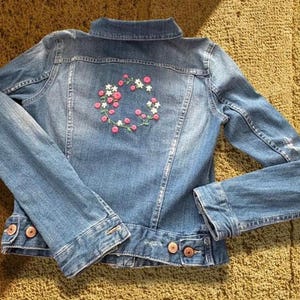 Puede incluir: Chaqueta vaquera azul claro con bordado floral en la espalda. El bordado presenta flores rosas y blancas dispuestas en un patrón circular. La chaqueta tiene botones y bolsillos.