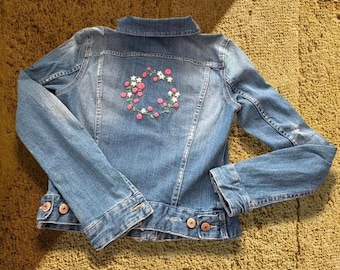 May include: A light blue denim jacket with a floral embroidery design on the back. The embroidery features pink and white flowers arranged in a circular pattern. The jacket has button closures and pockets.