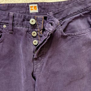 Puede incluir: Primer plano de unos pantalones vaqueros morados con botones plateados y un peque&ntilde;o bolsillo. Los pantalones tienen bragueta con botones y una peque&ntilde;a etiqueta con el logotipo naranja y blanco. La tela tiene un aspecto texturizado.
