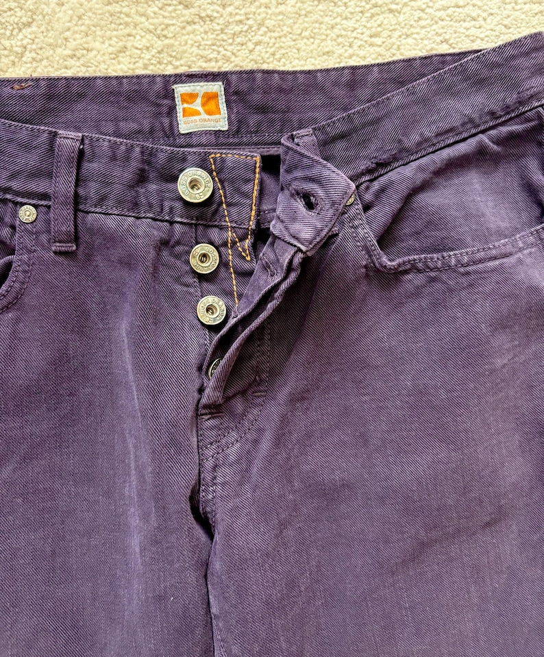 Puede incluir: Primer plano de unos pantalones vaqueros morados con botones plateados y un peque&ntilde;o bolsillo. Los pantalones tienen bragueta con botones y una peque&ntilde;a etiqueta con el logotipo naranja y blanco. La tela tiene un aspecto texturizado.
