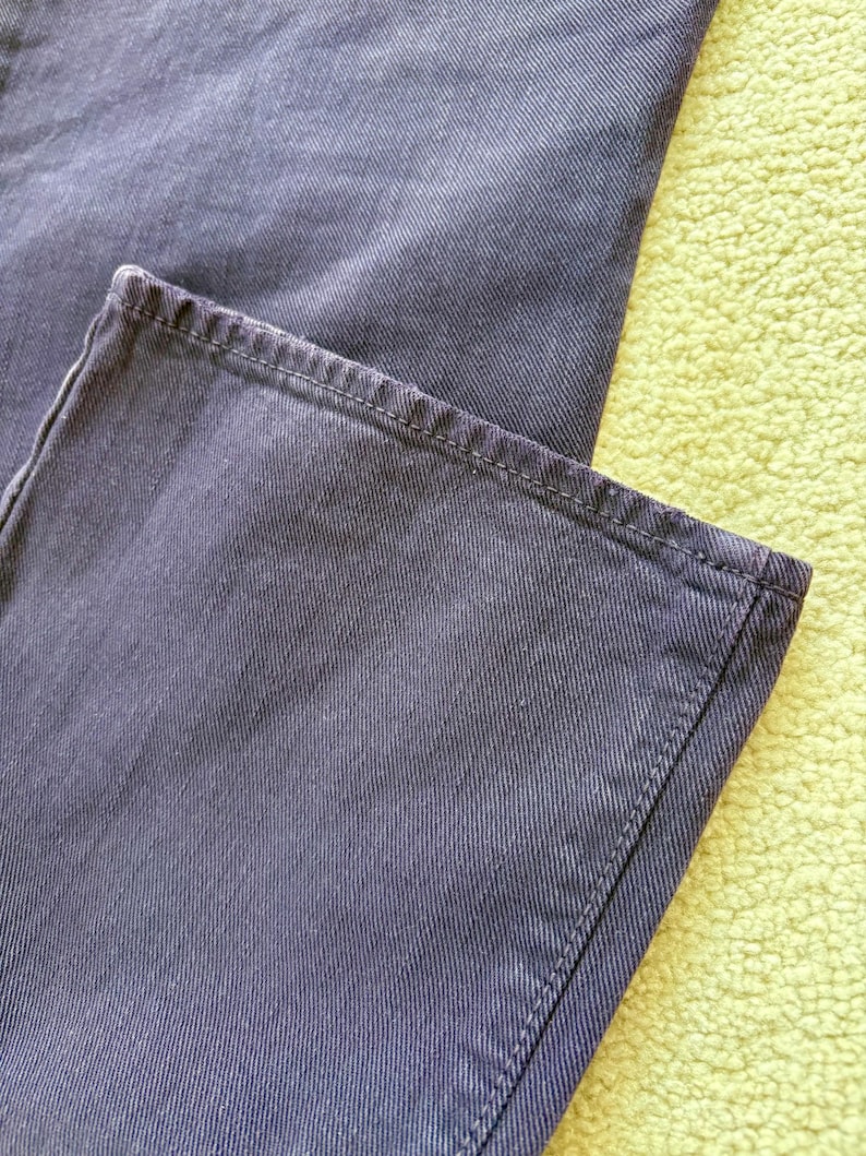 Puede incluir: Primer plano de una secci&oacute;n de vaqueros azul oscuro. La tela tiene una apariencia texturizada con l&iacute;neas diagonales visibles. El dobladillo est&aacute; cuidadosamente cosido con un hilo en contraste. Los vaqueros est&aacute;n sobre una superficie verde claro.