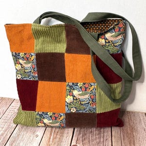 Może przedstawiać: Torba typu tote patchwork w różnych kolorach, w tym pomarańczowym, zielonym, brązowym i bordowym. Torba ma kwiatowy wzór z ptakami i truskawkami. Paski są w kolorze oliwkowym.