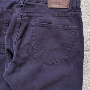 Puede incluir: Primer plano de unos vaqueros de mezclilla morados oscuros. Los vaqueros presentan un dise&ntilde;o cl&aacute;sico de cinco bolsillos, con un parche de cuero en la cintura que dice "BOSS ORANGE". El bolsillo trasero tiene un dise&ntilde;o cosido distintivo. La tela parece ser una mezclilla de peso medio.