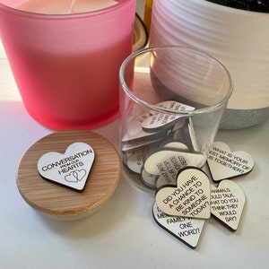 Puede incluir: Un tarro de vidrio lleno de peque&ntilde;os corazones de madera, cada uno con un iniciador de conversaci&oacute;n diferente escrito en &eacute;l. El tarro est&aacute; sentado en una mesa junto a una vela rosa y una planta en maceta. La tapa del tarro tiene un coraz&oacute;n de madera con las palabras "Conversation from our hearts" escritas en &eacute;l.