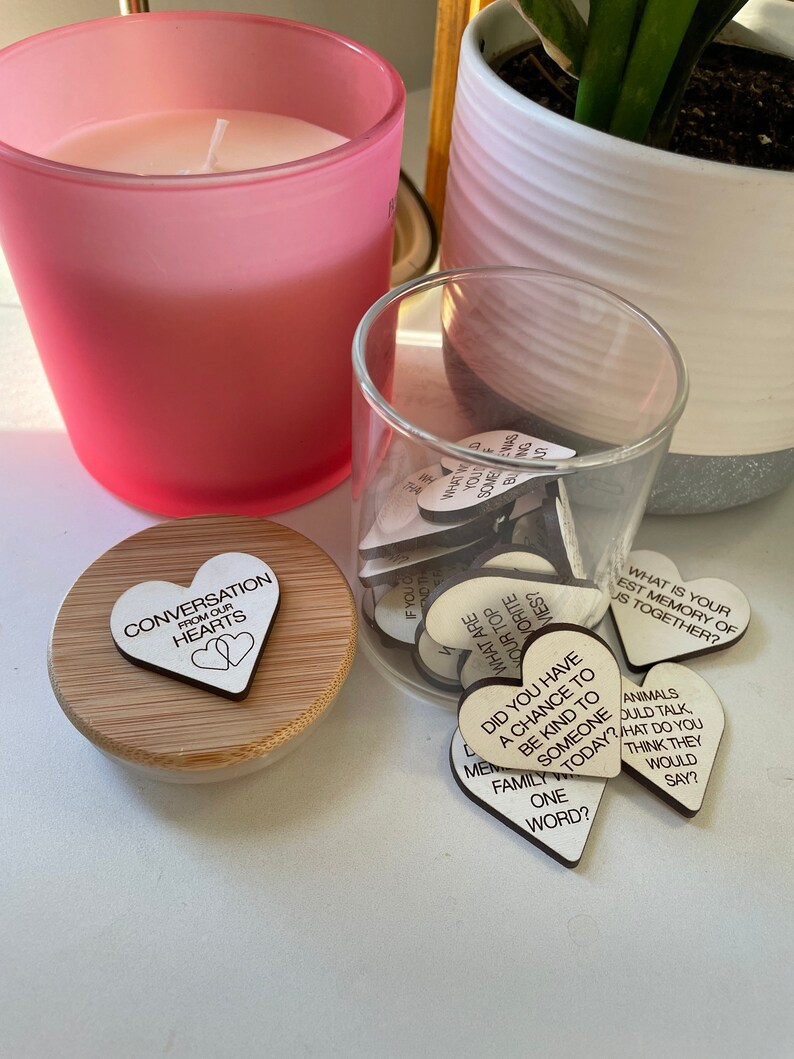 Puede incluir: Un tarro de vidrio lleno de peque&ntilde;os corazones de madera, cada uno con un iniciador de conversaci&oacute;n diferente escrito en &eacute;l. El tarro est&aacute; sentado en una mesa junto a una vela rosa y una planta en maceta. La tapa del tarro tiene un coraz&oacute;n de madera con las palabras "Conversation from our hearts" escritas en &eacute;l.