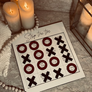 Puede incluir: Un juego de mesa blanco con las palabras "Strip Tic Toe" impresas en &eacute;l. El tablero de juego tiene nueve c&iacute;rculos con diferentes desaf&iacute;os escritos en ellos. Los c&iacute;rculos est&aacute;n rodeados de X negras.