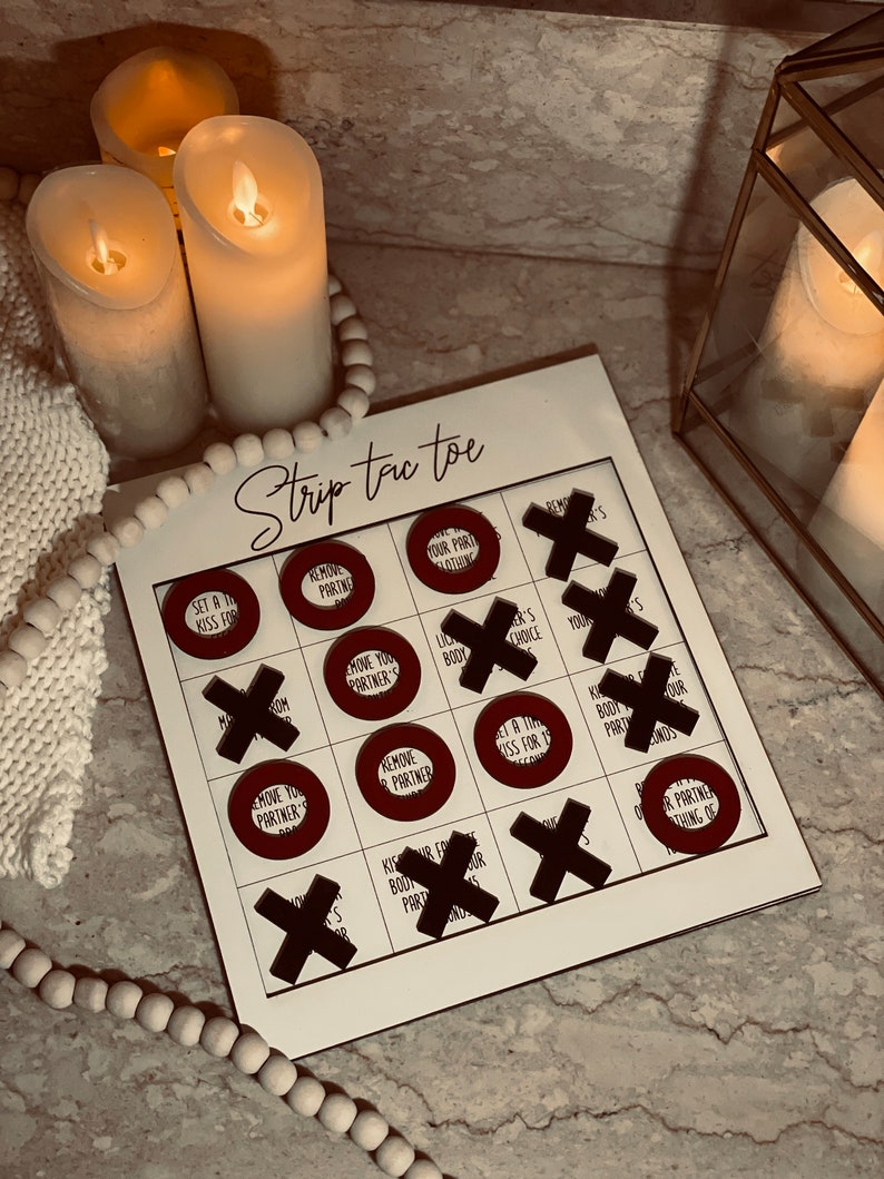 Puede incluir: Un juego de mesa blanco con las palabras "Strip Tic Toe" impresas en &eacute;l. El tablero de juego tiene nueve c&iacute;rculos con diferentes desaf&iacute;os escritos en ellos. Los c&iacute;rculos est&aacute;n rodeados de X negras.