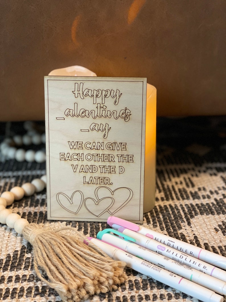 Puede incluir: Un letrero de madera con el texto "Happy Valentines-ay WE CAN GIVE EACH OTHER THE V AND THE D LATER." El letrero tiene tres corazones dibujados en &eacute;l.