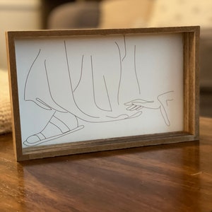 May include: A framed black and white line drawing of a person's foot wearing a sandal, with a hand reaching out from the bottom of the image.