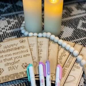 Puede incluir: Un conjunto de ocho tarjetas de madera para el D&iacute;a de San Valent&iacute;n con el texto "Happy Valentine's" y "Let me remind you that you are single" en una tarjeta. Las tarjetas est&aacute;n apiladas una encima de la otra y est&aacute;n rodeadas de marcadores blancos.