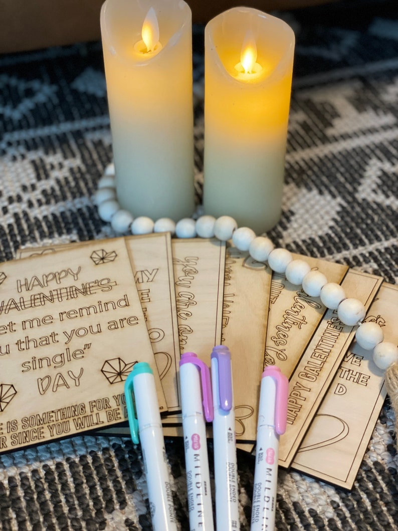 Puede incluir: Un conjunto de ocho tarjetas de madera para el D&iacute;a de San Valent&iacute;n con el texto "Happy Valentine's" y "Let me remind you that you are single" en una tarjeta. Las tarjetas est&aacute;n apiladas una encima de la otra y est&aacute;n rodeadas de marcadores blancos.