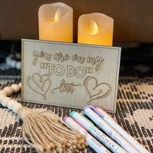 Puede incluir: Un letrero de madera con el texto "you are on my "TO DO" list" y dos corazones. El letrero est&aacute; sobre una mesa con dos velas encendidas detr&aacute;s. Tambi&eacute;n hay tres rotuladores de colores pastel y un borla marr&oacute;n sobre la mesa.