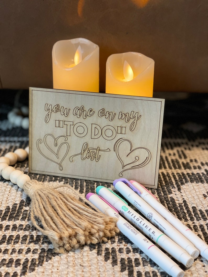 Puede incluir: Un letrero de madera con el texto "you are on my "TO DO" list" y dos corazones. El letrero est&aacute; sobre una mesa con dos velas encendidas detr&aacute;s. Tambi&eacute;n hay tres rotuladores de colores pastel y un borla marr&oacute;n sobre la mesa.