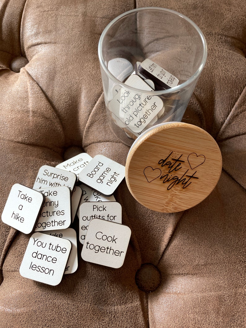Puede incluir: Un tarro de vidrio lleno de peque&ntilde;os cuadrados de madera con diferentes ideas de citas escritas en ellos. El tarro est&aacute; sentado en un sof&aacute; marr&oacute;n con una tapa de madera que dice "date night" con un coraz&oacute;n.