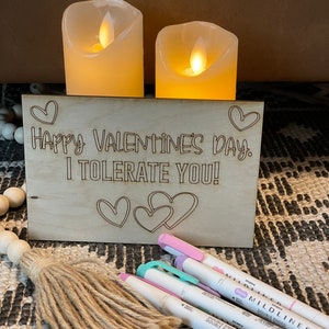 Puede incluir: Un letrero de madera con el texto "Happy Valentine's Day. I tolerate you!" El letrero est&aacute; decorado con corazones y est&aacute; sobre una mesa con dos velas blancas.
