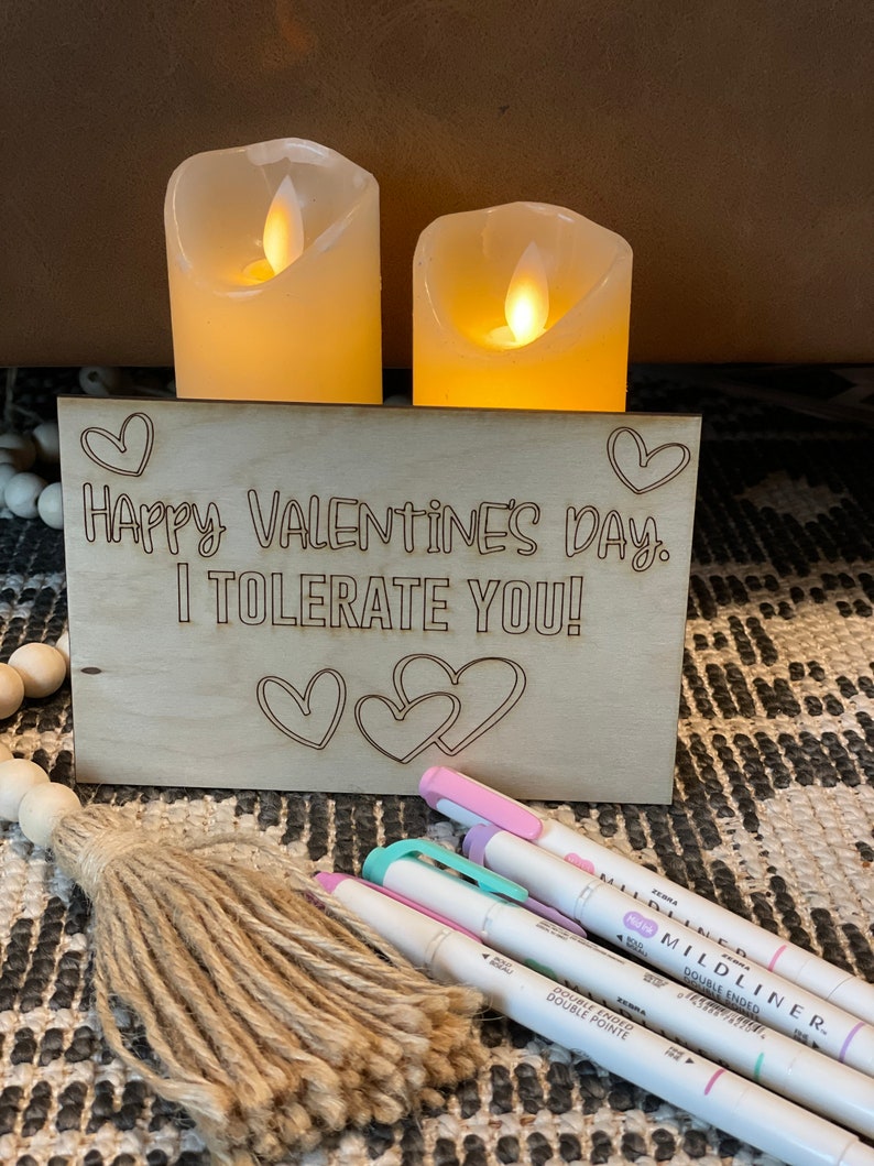 Puede incluir: Un letrero de madera con el texto "Happy Valentine's Day. I tolerate you!" El letrero est&aacute; decorado con corazones y est&aacute; sobre una mesa con dos velas blancas.