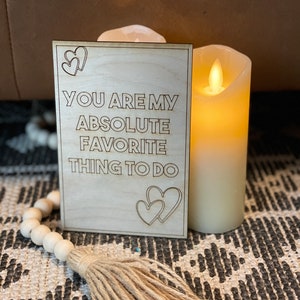 Puede incluir: Un letrero de madera con el texto "You are my absolute favorite thing to do" y dos formas de coraz&oacute;n a cada lado del texto. El letrero est&aacute; sobre una alfombra con una vela en el fondo.