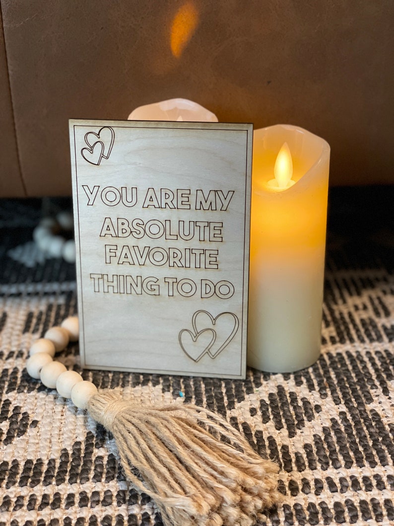 Puede incluir: Un letrero de madera con el texto "You are my absolute favorite thing to do" y dos formas de coraz&oacute;n a cada lado del texto. El letrero est&aacute; sobre una alfombra con una vela en el fondo.