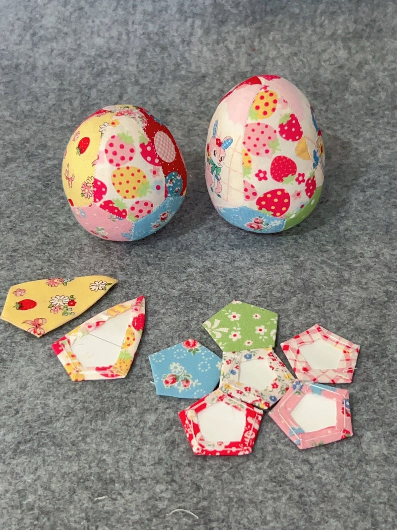 May include: Two fabric Easter eggs with patchwork patterns of strawberries, flowers, and a bunny. Several fabric pentagon shapes with white centres are scattered nearby. The eggs are primarily pink, red, yellow, and blue.