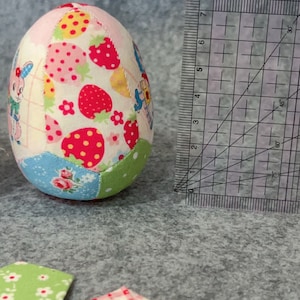 May include: A fabric Easter egg with a patchwork design. The egg features various fabric patterns, including strawberries, floral prints, and cartoon characters. A ruler is next to the egg, showing measurements in centimetres.