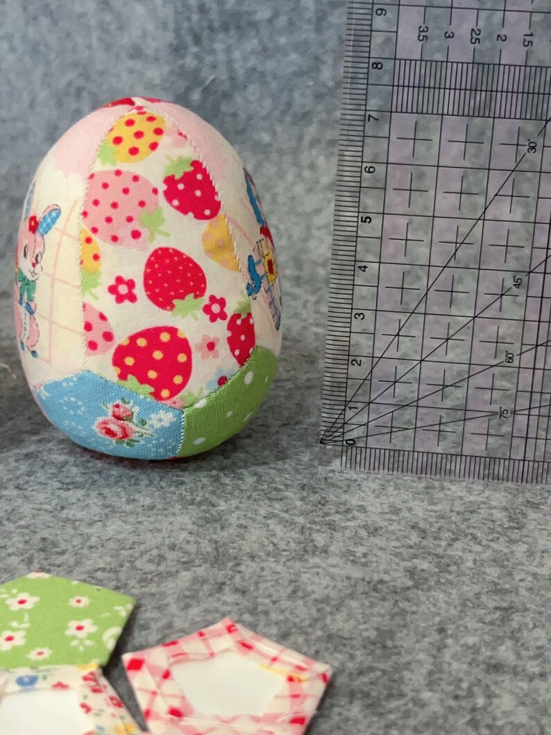 May include: A fabric Easter egg with a patchwork design. The egg features various fabric patterns, including strawberries, floral prints, and cartoon characters. A ruler is next to the egg, showing measurements in centimetres.