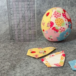 May include: A fabric egg with a patchwork design featuring strawberries and flowers. A clear ruler with centimetre markings is next to the egg. Two fabric pieces with floral patterns are in the foreground.