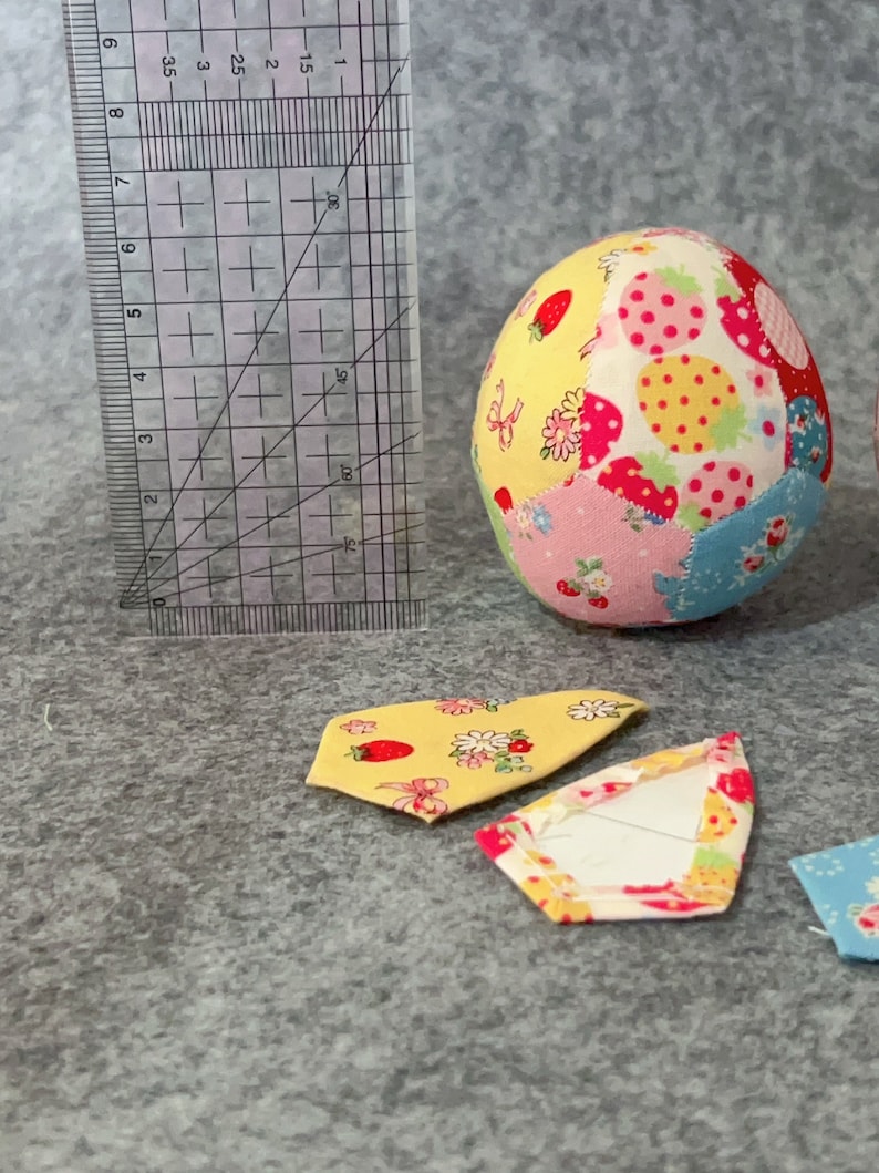 May include: A fabric egg with a patchwork design featuring strawberries and flowers. A clear ruler with centimetre markings is next to the egg. Two fabric pieces with floral patterns are in the foreground.