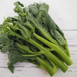 May include: A bunch of fresh green Chinese broccoli, also known as gai lan, with stems and leaves. The vegetable is arranged on a white wooden surface.