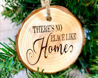 以下が含まれることがあります： 「THERE'S NO PLACE LIKE Home」の文字が黒で刻まれた、丸い木のクリスマスオーナメント。オーナメントは天然の木の輪切りで作られ、麻ひものループから吊り下げられています。オーナメントは緑のクリスマスツリーに飾られています。