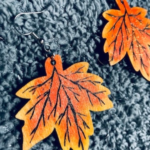 May include: A pair of wooden leaf earrings with a glittery orange and yellow finish. The leaves have black veins and are attached to silver hooks.