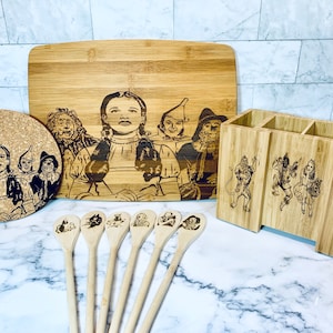 May include: A collection of kitchenware items with engraved images from The Wizard of Oz. Includes a cutting board, a coaster, a utensil holder, and wooden spoons. The items are made of bamboo and wood.