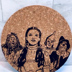 May include: A cork coaster with a black and white illustration of Dorothy and her friends from the Wizard of Oz. The characters are depicted in a close-up view, with Dorothy in the center, the Cowardly Lion on the left, the Tin Man on the right, and the Scarecrow in the back.