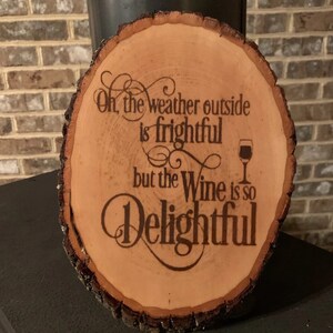 Könnte beinhalten: Ein Holzscheibe mit dem Text "Oh, das Wetter draußen ist furchtbar, aber der Wein ist so köstlich" und einem kleinen Bild eines Weinglases.