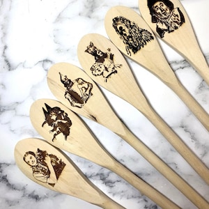 May include: Six wooden spoons featuring different characters from The Wizard of Oz, burned into the wood. The characters include Dorothy, the Scarecrow, the Tin Man, the Cowardly Lion, and the Wicked Witch of the West.