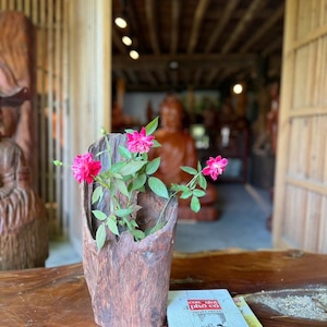 May include: A rustic wooden vase holds vibrant pink roses and green leaves. The vase sits on a wooden table next to a book with text. The background features a wooden structure and a statue.