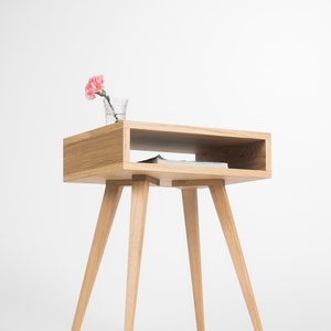 Peut inclure: Une table d'appoint en bois clair avec une étagère et quatre pieds. Une fleur rose dans un vase en verre est posée sur l'étagère supérieure.