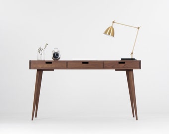May include: A dark wood desk with three drawers and a gold metal desk lamp. The desk has tapered legs and is sitting on a white surface.